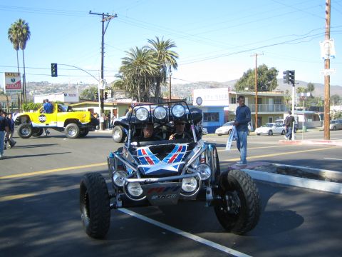 1990 Ford Bronco - blue and white off-road dune buggy with roof-mounted lights stopped on a sunny street, with a yellow off-road truck and palm trees in the b…