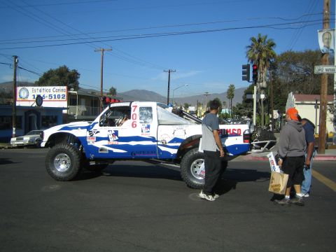 1990 Ford Bronco - white and blue off-road race truck numbered 706 parked on a street with bystanders nearby and hills in the background.