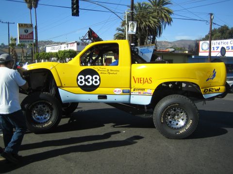 1990 Ford Bronco - yellow lifted off-road race truck numbered 838 with Viejas and DRW sponsor decals, a man walking alongside it on a sunny street.