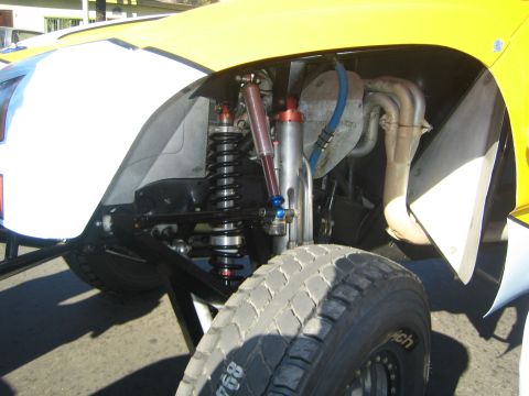 1990 Ford Bronco - Close-up of an off-road race truck's rear suspension showing a black coilover and red bypass shocks behind a tire under a yellow fender.