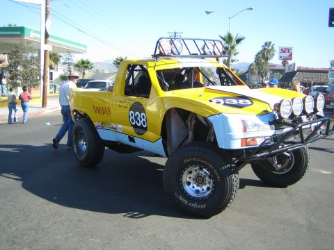 1990 Ford Bronco - yellow lifted off-road race truck numbered 838 with Viejas decals and roof lights parked on a street near a Pemex station with spectators.