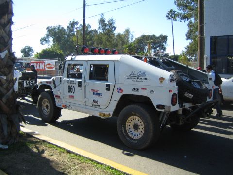 1990 Ford Bronco - white Hummer H1 off-road race vehicle numbered 860 with roof-mounted lights and a rear spare tire parked on a street lined with palm trees.