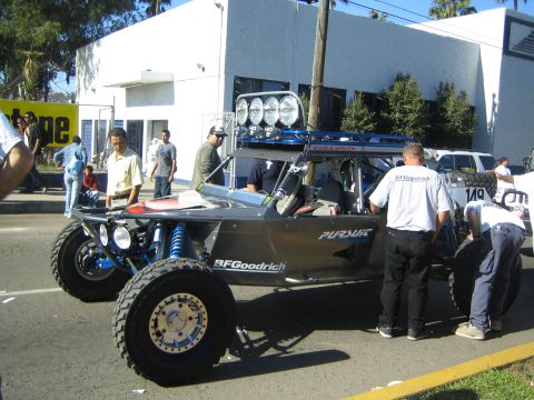 1990 Ford Bronco - silver multi-seat off-road buggy with BFGoodrich and Pursuit decals and roof-mounted lights, with crew members working on it on a street.