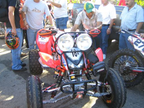 1990 Ford Bronco - red and black off-road racing ATV quad numbered 16 with dual headlights and roll cage, surrounded by men inspecting it on dirt ground.