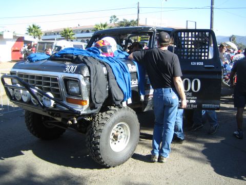 1990 Ford Bronco - black vintage Ford off-road race truck numbered 300 with a front light bar and roll cage, a person in black standing beside it at a pit are…