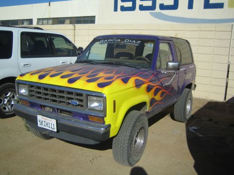 1990 Ford Bronco - two-door Ford SUV painted purple with yellow and orange flame graphics and a California license plate, parked beside a building.