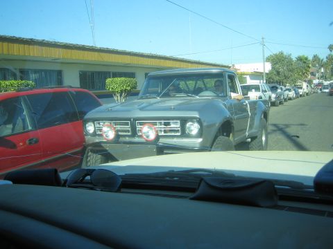 1990 Ford Bronco - gray vintage pickup truck with round dual headlights driving on a street, photographed over the dashboard of another vehicle.