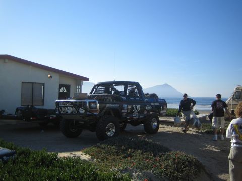 1990 Ford Bronco - black off-road race truck numbered 300 parked beside a building near the beach, with people unloading gear and an RV in the background.