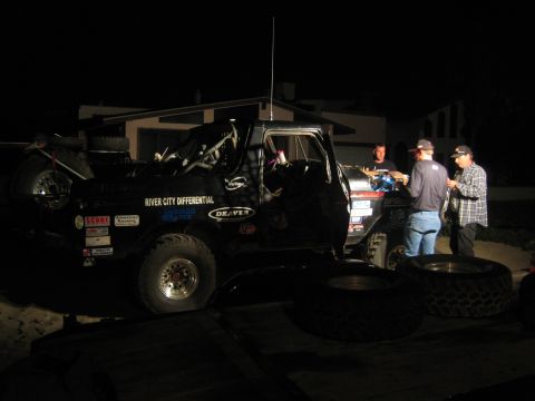 1990 Ford Bronco - heavily decaled black off-road race truck sits on a trailer at night as three men work nearby among stacked spare tires.