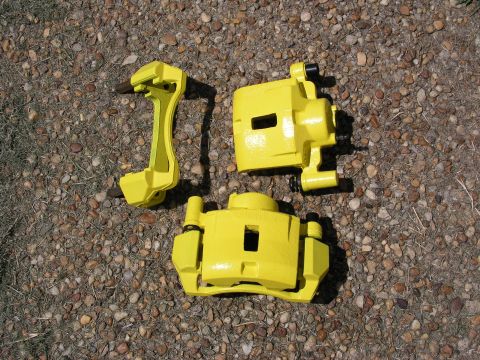 1996 Ford Probe - A pair of bright yellow brake calipers and their mounting bracket are displayed on a bed of small stones and dirt.