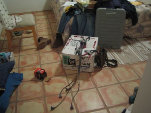 1990 Ford Bronco - tiled room strewn with gear: a battery charger box with cables, work boots, folded clothing and a gray hard-shell case.