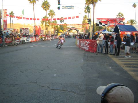 1990 Ford Bronco - dirt-bike rider rides up a city street toward a banner-draped finish arch flanked by Coca-Cola banners and crowds.