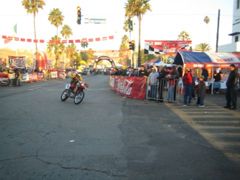 1990 Ford Bronco - motorcycle racer rides down a palm-lined street past Coca-Cola banners, vendor tents and a row of spectators.