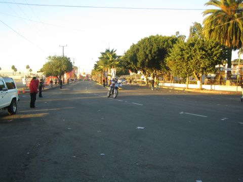 1990 Ford Bronco - lone motorcyclist rides down a wide, mostly empty street in early light, with palm trees and scattered bystanders.