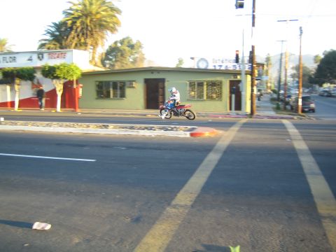 1990 Ford Bronco - dirt-bike rider crosses a paved intersection in front of a green roadside building bearing Spanish-language signs.