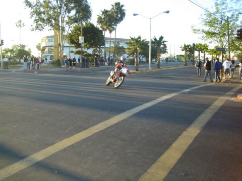 1990 Ford Bronco - dirt-bike rider moves along a broad paved road in low morning light, casting long shadows past palms and onlookers.