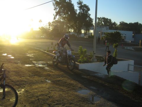 1990 Ford Bronco - dirt-bike rider pauses beside a landscaped median in bright sunrise glare, near a parked bicycle and a passerby.