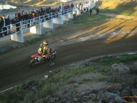 1990 Ford Bronco - motorcycle racer powers along a dirt course below a packed crowd watching from a white fence on the embankment.