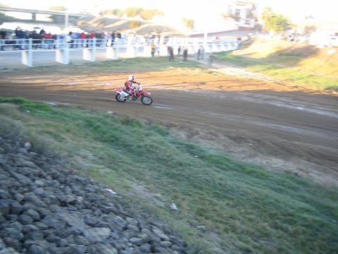 1990 Ford Bronco - dirt-bike rider rides a wide dirt course near a concrete bridge as spectators line the rail along the slope.