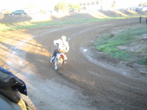 1990 Ford Bronco - dirt-bike rider rounds a banked dirt turn, with parked vehicles and an arched building visible in the background.