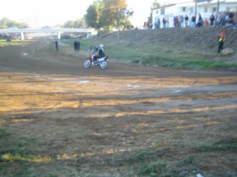 1990 Ford Bronco - dirt-bike rider crosses an open dirt track near a bridge as spectators gather by a white building on the rise.
