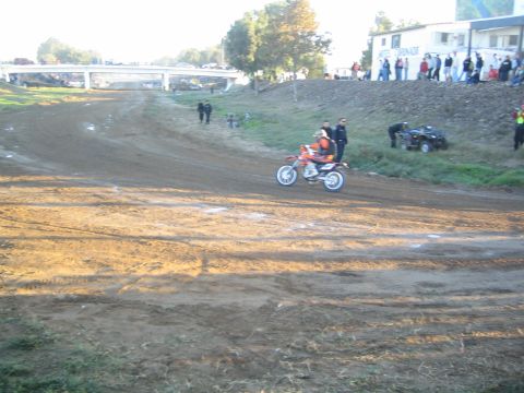1990 Ford Bronco - rider on an orange dirt bike moves down a dirt course past officials, a parked ATV and spectators on the bank.