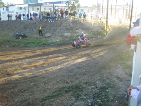 1990 Ford Bronco - red racing ATV kicks up dust through a dirt turn below a crowd lined along the bank near a motel building.