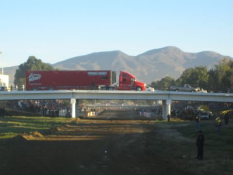 1990 Ford Bronco - red semi-truck and trailer cross an overpass spanning a dirt race course, with mountains rising in the background.
