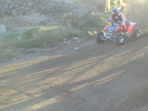 1990 Ford Bronco - red racing ATV and helmeted rider speed along a dirt track beside a grassy, brush-covered embankment.
