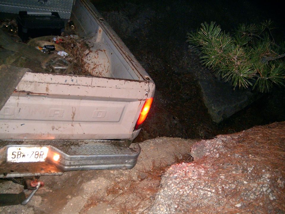 Rear view of a light-colored Ford pickup truck, license plate SB-78R, with the tailgate down and bed loaded with debris. Off-road terrain is visible