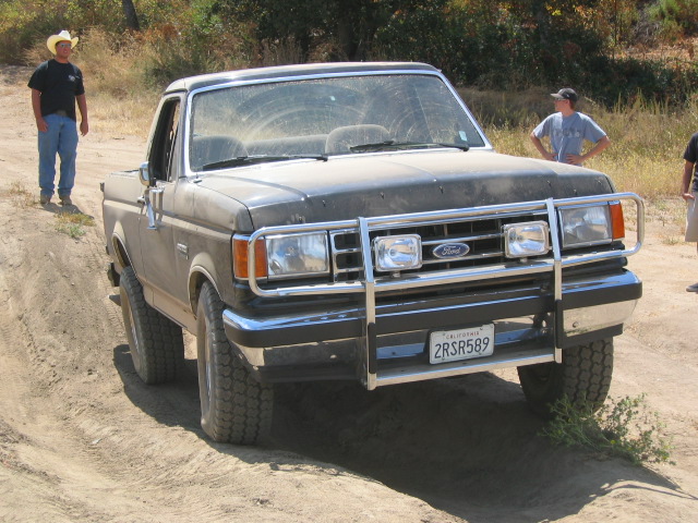 Dusty dark Ford Bronco with chrome brush guard flexing on a dirt trail, front 3/4 view