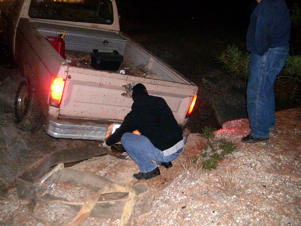 Two people are working on the rear of a pickup truck. One person is crouching down, and another is standing nearby