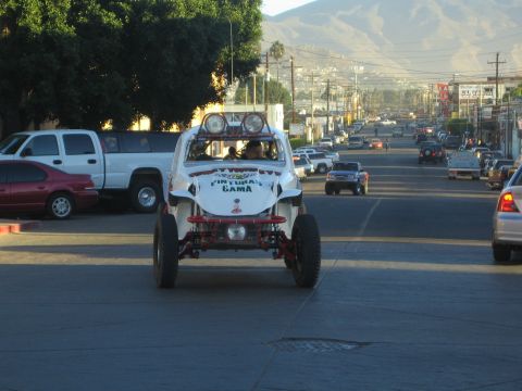 1990 Ford Bronco - white open-cockpit off-road buggy drives away down a town street lined with parked cars, trucks and power poles.