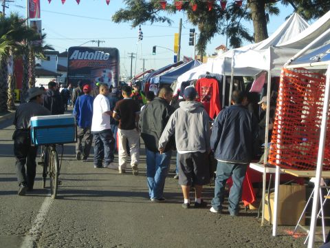 1990 Ford Bronco - People crowd a street market lined with vendor tents and orange fencing, with a large Autolite trailer in the background.