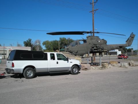 1990 Ford Bronco - A white pickup truck with a black camper shell is parked next to a military helicopter mounted on a pole.