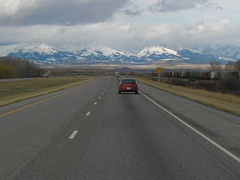 1990 Ford Bronco - A red sedan travels down a highway, with a train and snowy mountains visible.