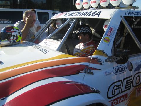 1990 Ford Bronco - driver in a checkered helmet sits in a white Yokohama-marked desert race truck while a man leans against the door.