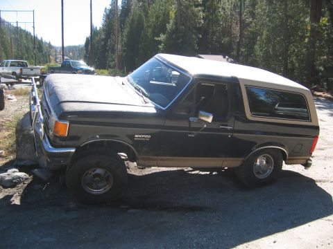 1990 Ford Bronco - Side view of a 1990 Ford Bronco parked off-road, with other vehicles in the distance.