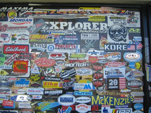 1990 Ford Bronco - dense collage of off-road racing and brand decals including Edelbrock, KORE and McKenzie's covers a window.