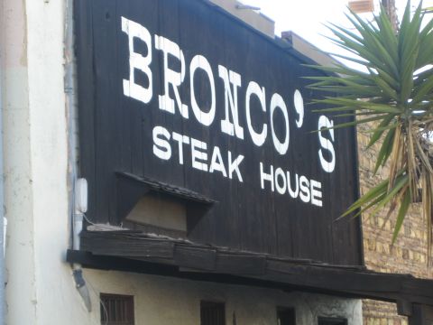 1990 Ford Bronco - weathered black wooden sign with white lettering reads BRONCO'S STEAK HOUSE beside a palm-like plant.