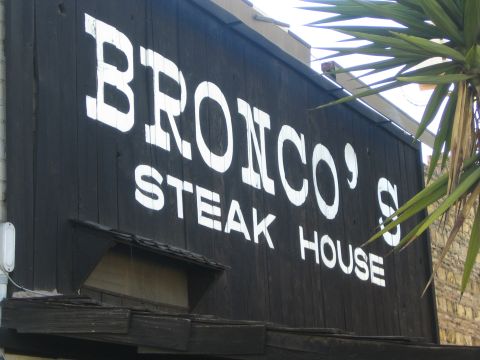 1990 Ford Bronco - close view of a weathered black wooden BRONCO'S STEAK HOUSE sign with white lettering and overhanging palm fronds.