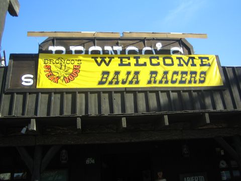 1990 Ford Bronco - yellow banner reading WELCOME BAJA RACERS hangs across the front of the dark wooden Bronco's Steak House building.