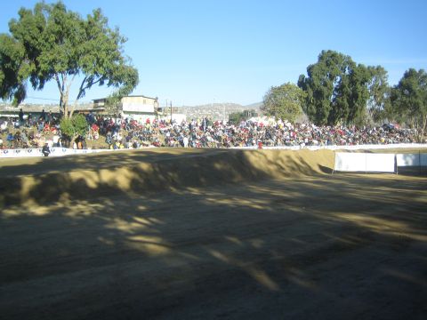 1990 Ford Bronco - Spectators line a tree-backed dirt hillside under a clear blue sky, viewed across a shadowed empty dirt track.