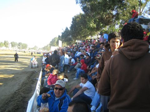 1990 Ford Bronco - Spectators crowd a dirt embankment along a track, some seated on a barrier, with a man in brown in the foreground.