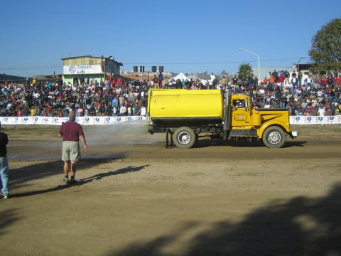 1990 Ford Bronco - yellow tanker truck sprays water on a dirt track to control dust in front of a packed grandstand crowd.