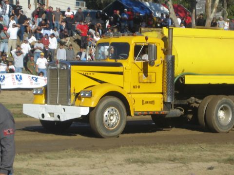 1990 Ford Bronco - yellow Kenworth tanker truck with chrome grille drives along a dirt track as a crowd watches behind a banner.