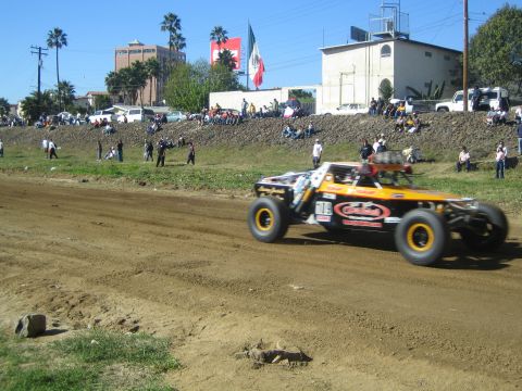 1990 Ford Bronco - orange and white off-road trophy truck races on a dirt course with spectators and buildings behind under a Mexican flag.