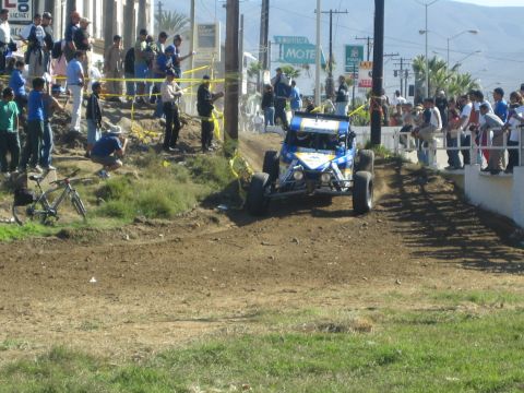 1990 Ford Bronco - blue single-seat off-road buggy climbs a dirt course lined with spectators near a street and motel sign.
