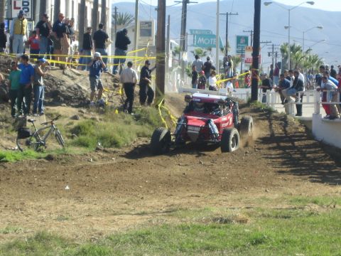 1990 Ford Bronco - red off-road buggy navigates a dirt course as spectators and tape barriers line the edge near a street.