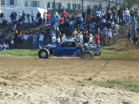 1990 Ford Bronco - blue off-road race truck numbered 122 kicks up dirt on a course in front of a large standing crowd.